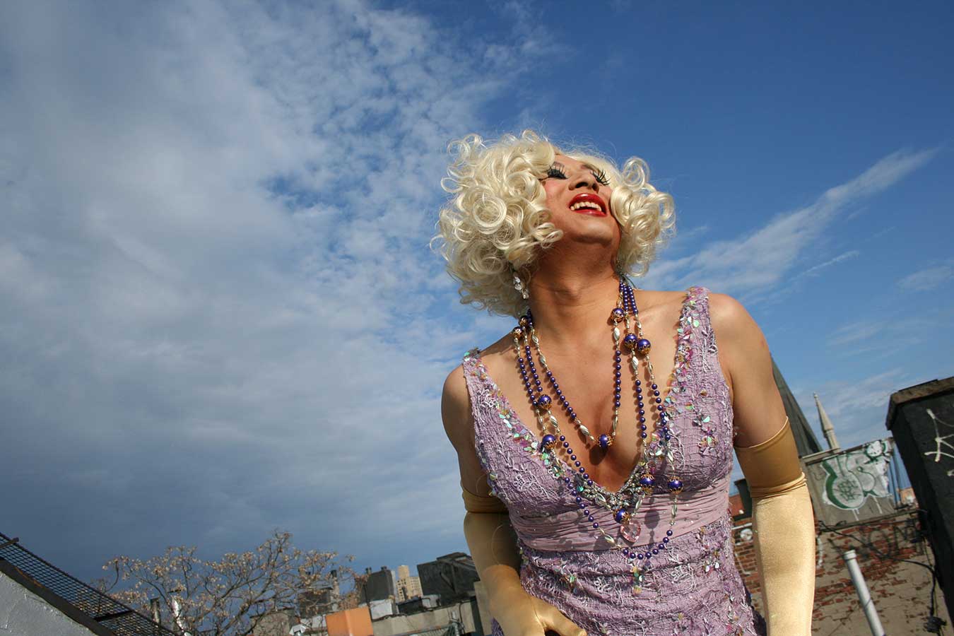 An East Asian drag queen photographed from a crouching position, so that they're looking into the sun behind the photographer, with their Marilyn Monroe curls juxtaposed against a blue sky with light clouds.