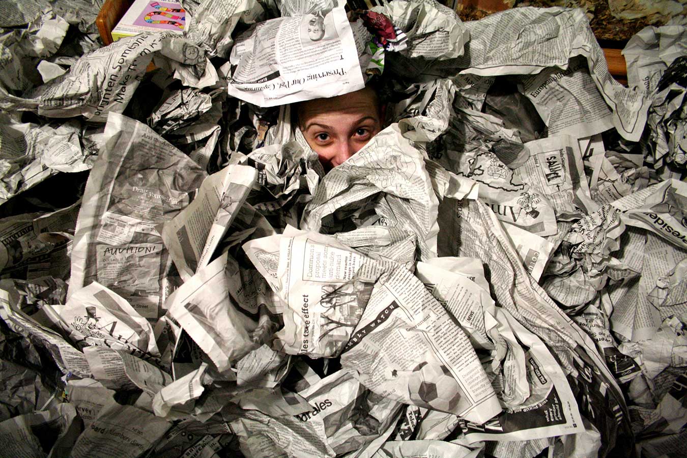 A white college student peers out from a pile of crumpled newspaper, only her forehead, eyes, and nose visible.
